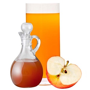apple cider vinegar in a carafe with spout, in a tall cylinder container with half an apple at bottom right.