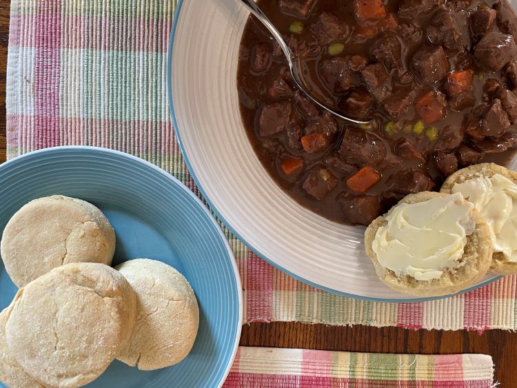 Beef stew with spoon on plate with buttered gluten free biscuit.  Gluten free biscuits on a side plate.