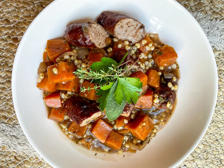 braised sausages with carrots and sorghum in a white bowl garnished with fresh herbs.