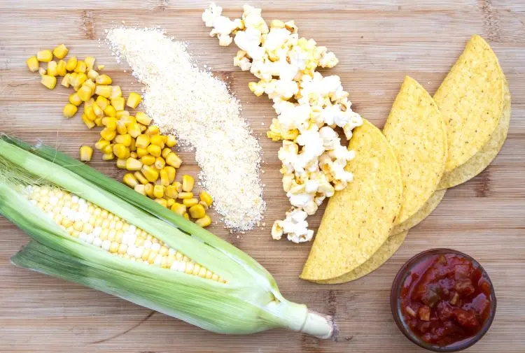 overhead shot of corn on the cob, corn flour, popcorn, corn meal and corn tortillas on a wooden board.
