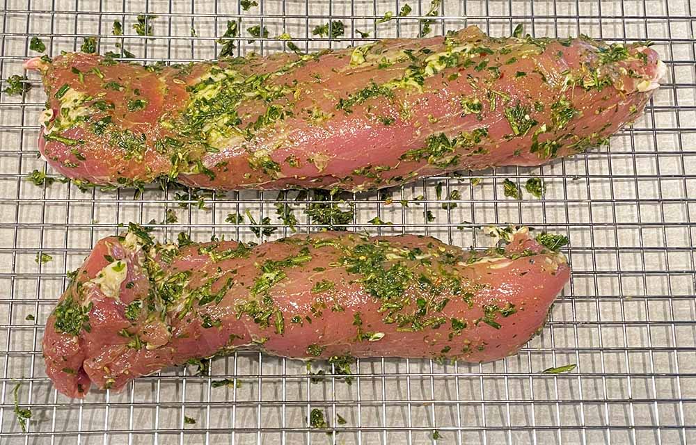 two raw pork tenderloins covered in herb paste on a metal rack.