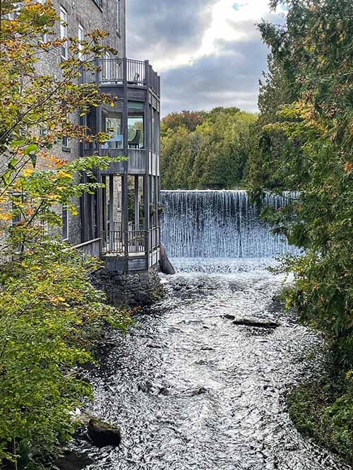 Millcroft Inn Caledon Ontario Canada.  Photo of waterfall and sparkling river alongside rustic inn and green foliage.