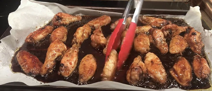 baked, sauce covered chicken wings on parchment lined baking sheet and tongs turning the wings in the sauce. baked, sauce covered chicken wings on parchment lined baking sheet and tongs turning the wings in the sauce.